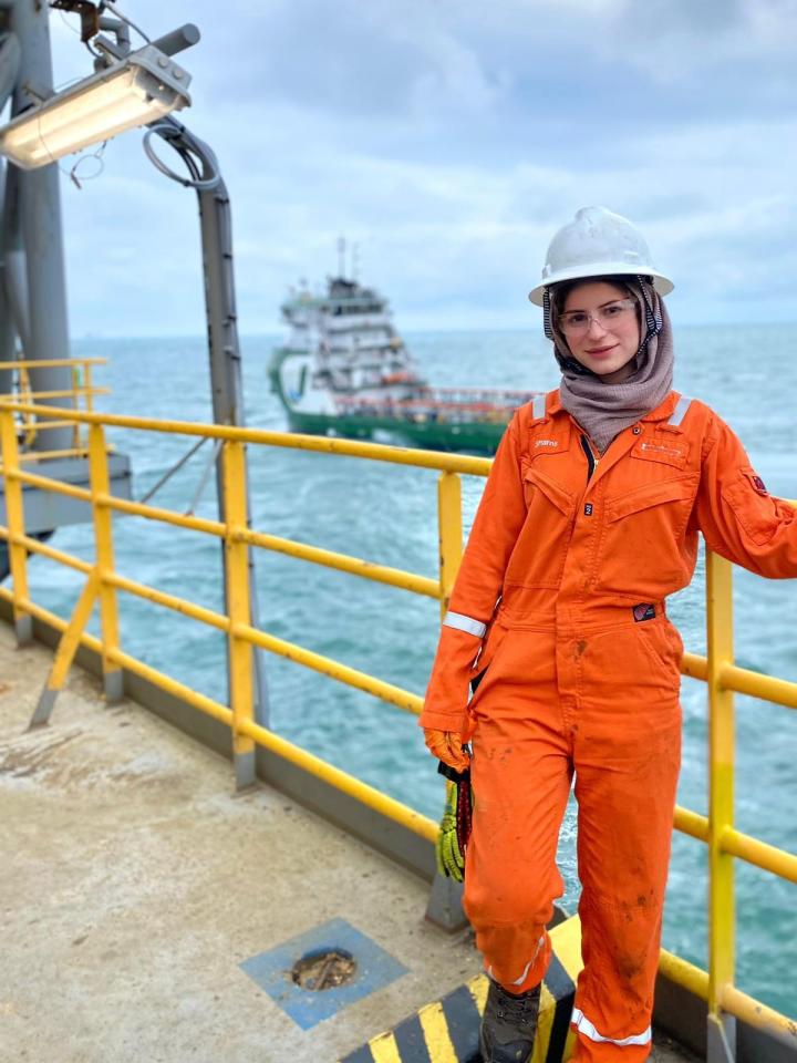 a woman wearing an orange jumpsuit and a hard hat stands on a deck