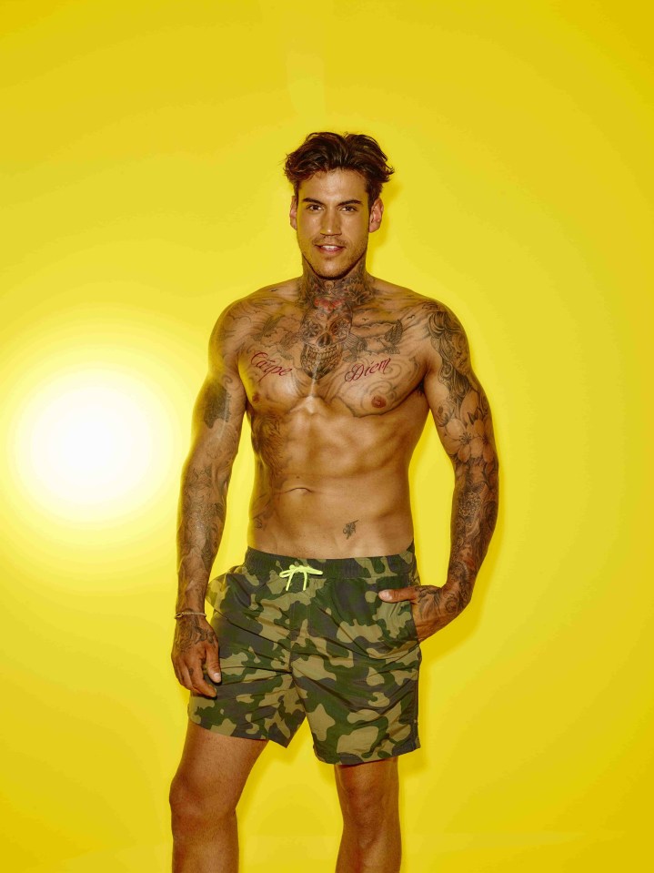 A muscular man with tattoos wearing camouflage swim trunks against a yellow background.