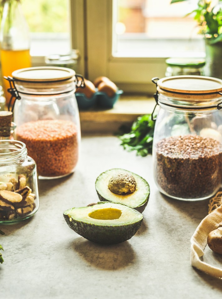 Sliced avocado, glass jars with lentils and nuts at kitchen table with various ingredients at window background with natural light. Healthy cooking at home. Front view.