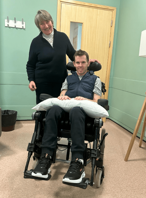 a woman stands next to a man in a wheelchair