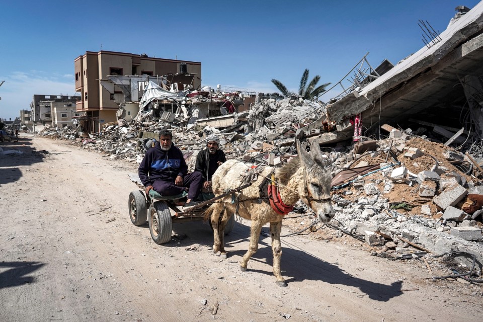 Two men sit in a donkey-drawn cart moving past the rubble