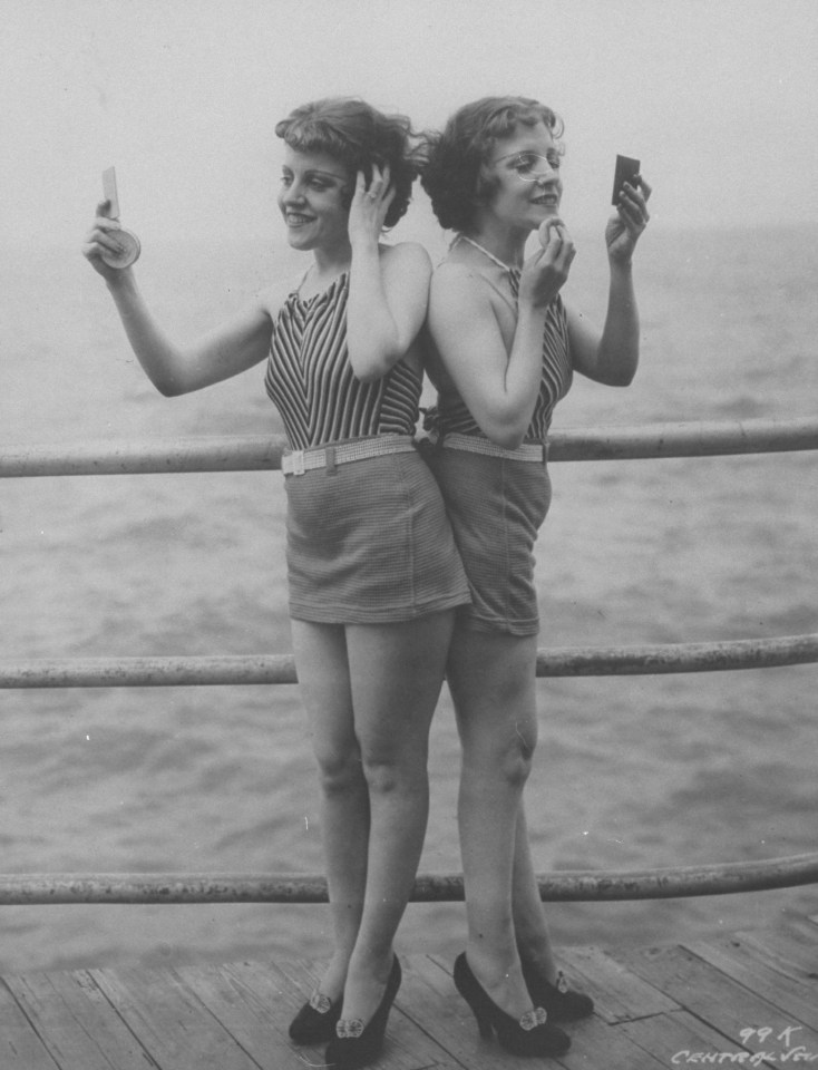 Violet and Daisy Hilton stand back to back looking in hand mirrors and powdering faces on pier at Atlantic City in 1946