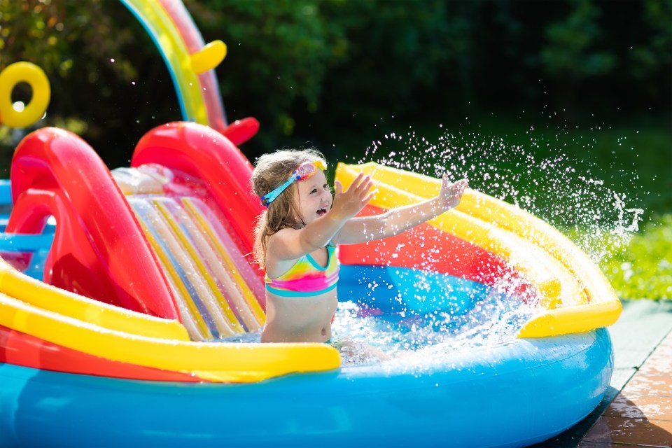 Child playing in inflatable baby pool. Kids swim, slide and splash in colorful garden play center. Happy little girl sliding and swimming with water toys on hot summer day. Family outdoor fun.