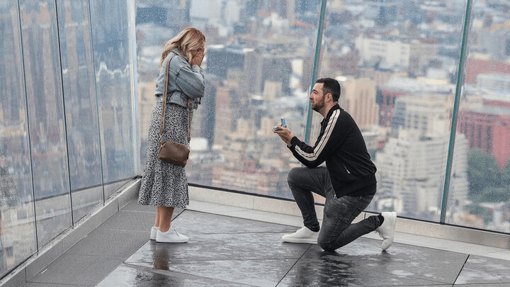 A man proposing to a woman on a rooftop overlooking a city.