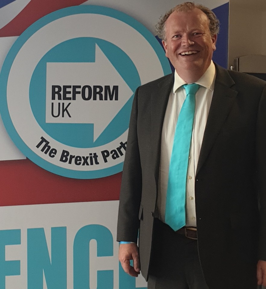 a man in a suit stands in front of a reform uk sign