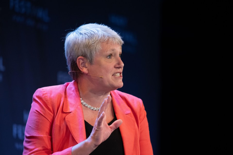 a woman wearing a pink jacket and a pearl necklace speaks into a microphone