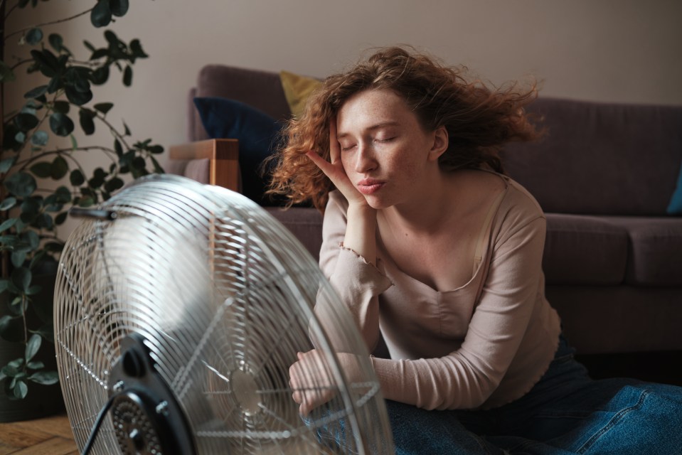 Suffering from summer heat at home, exhausted young woman sitting near electric fan enjoying fresh air, cooling down during heatwave