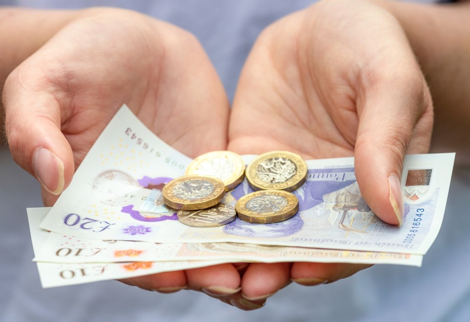 Macro image of a woman's hands as she holds out British ten and twenty pound notes, with pound coins and a twenty pence piece.