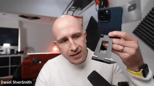 a bald man is holding a cell phone in front of a microphone with daniel shensmith written below him