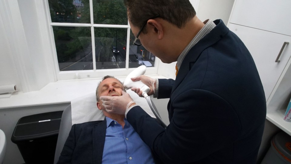 a man in a blue shirt is laying on a bed while a man in a suit holds a device over his face