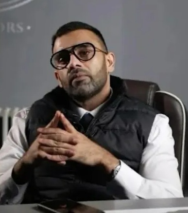 a man wearing glasses and a vest is sitting at a table with his hands folded