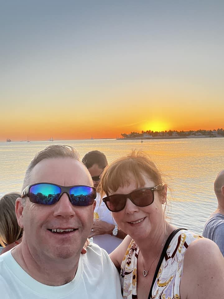 a man and woman wearing sunglasses pose for a picture in front of a sunset over the ocean