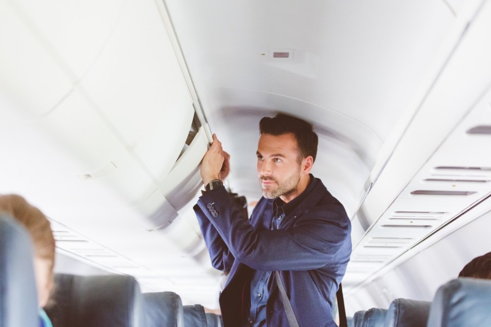 Mature man travelling by flight storing handbag. Airplane passenger in the cabin storing hand luggage in the overhead locker.