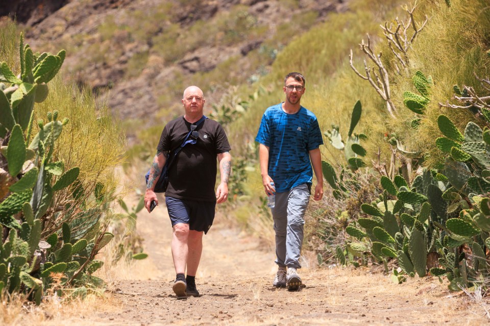 two men walking down a dirt path with cactus in the background