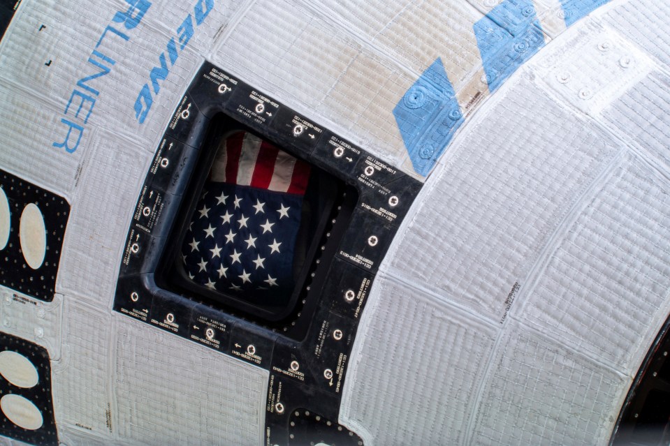 A U.S. flag is displayed from a window of the Boeing Starliner spacecraft docked to the ISS