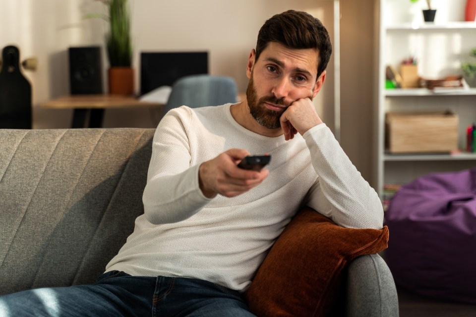 Photo of young bored man sitting on couch at home and changing TV channels with disinterest. Stock photo