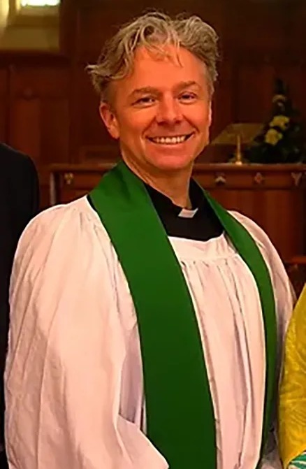 a man in a white robe and green sash smiles in front of a stained glass window