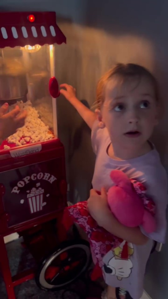 a little girl is holding a stuffed animal in front of a popcorn machine