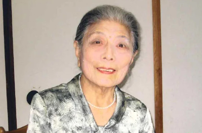 an elderly woman wearing a pearl necklace is sitting in a chair and smiling .