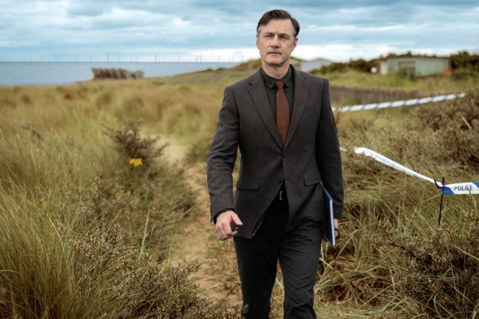 a man in a suit and tie walks through a field with a police tape behind him