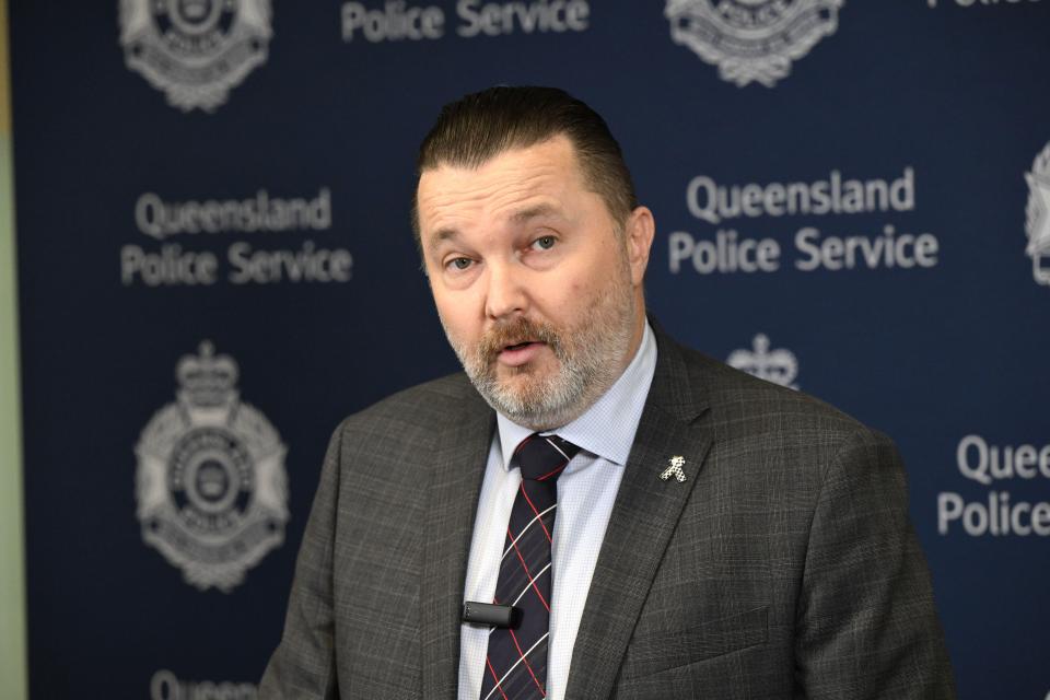 a man in a suit and tie stands in front of a queensland police service sign