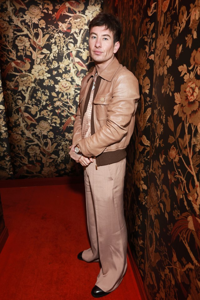 a man in a tan leather jacket stands in front of a wall with floral wallpaper