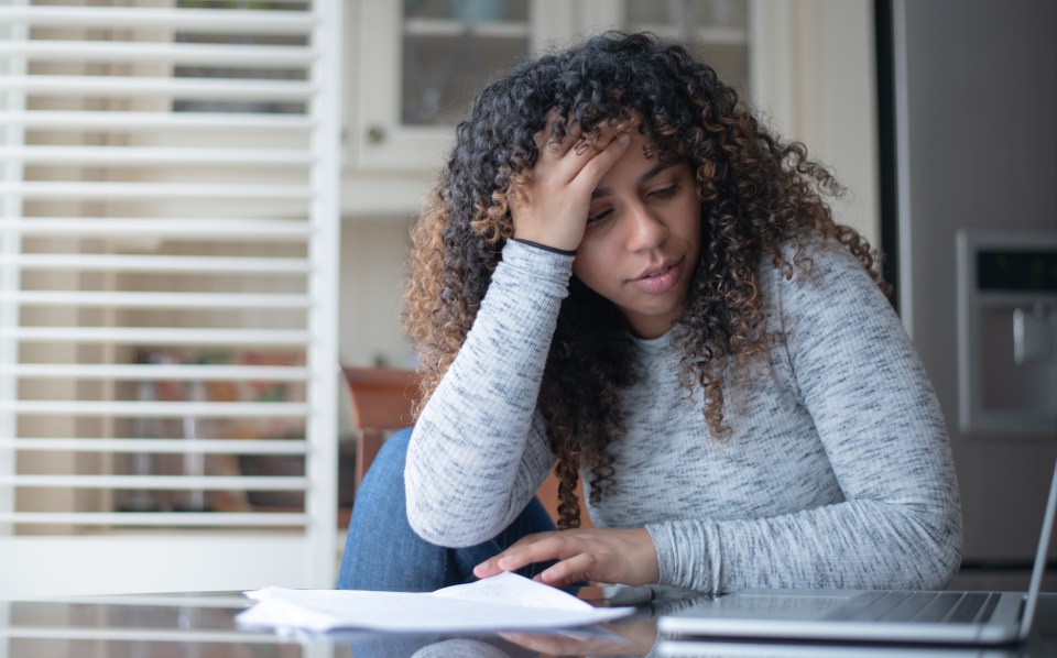 A mixed race woman sit at a table with one knee bent up on her chair.  She has her head resting in her hand with her elbow on the table and is looking frustrated.  Shed is wearing casual clothing.