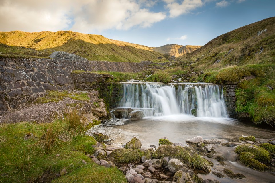 a waterfall is surrounded by grass and rocks with mountains in the background