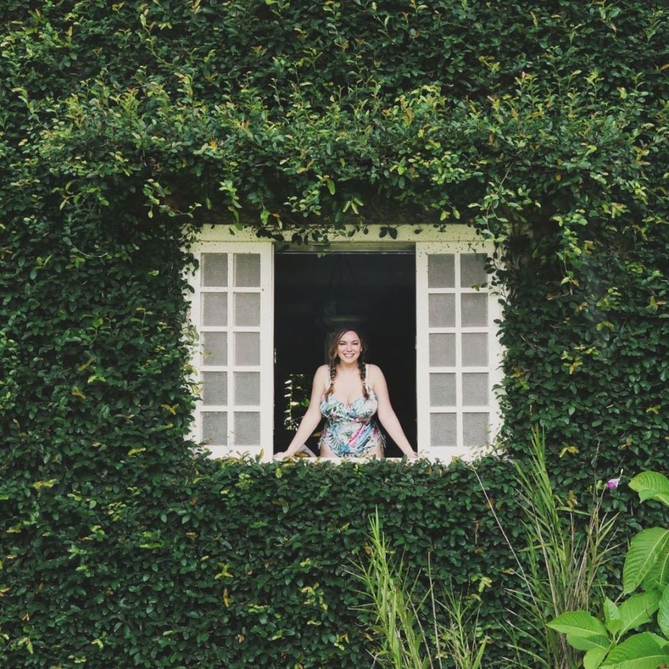 a woman in a bathing suit stands in a window surrounded by greenery