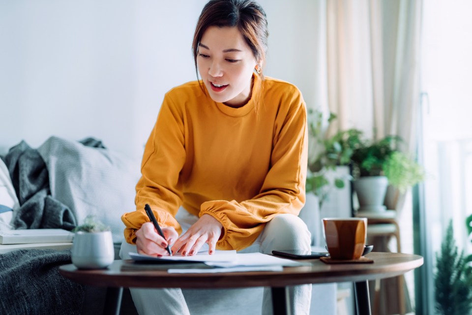 Young Asian woman holding a pen and signing paperwork in the living room at home. Deal concept