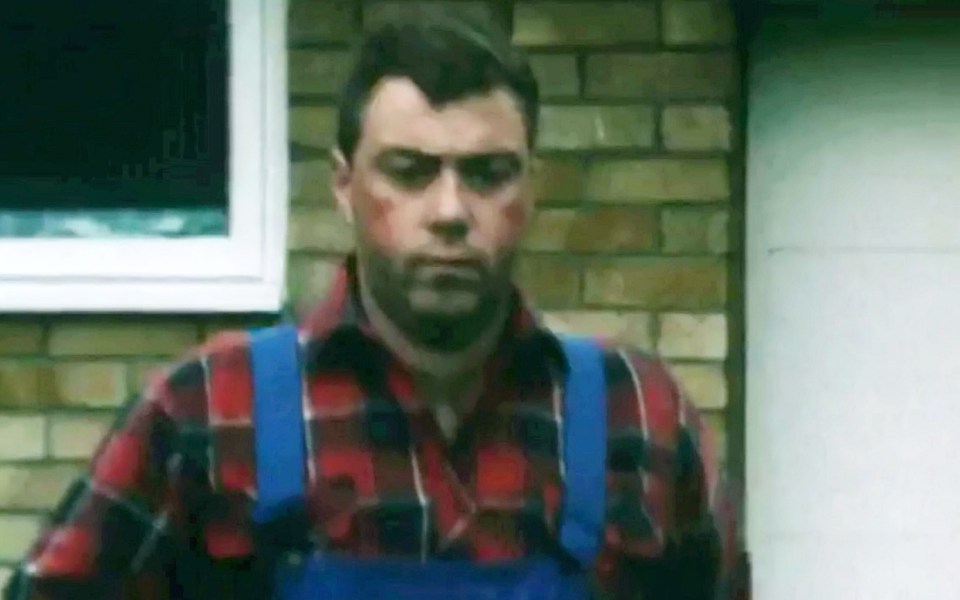 a man in a plaid shirt and blue overalls is standing in front of a brick wall .