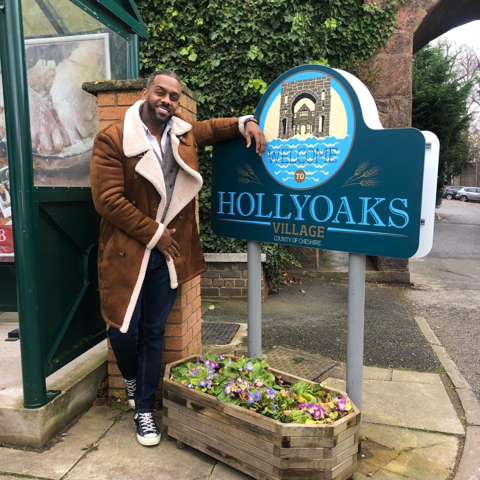 a man stands in front of a hollyoaks village sign