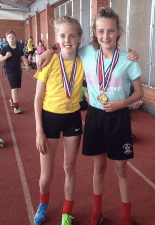 Two girls with medals around their necks stand on a running track.