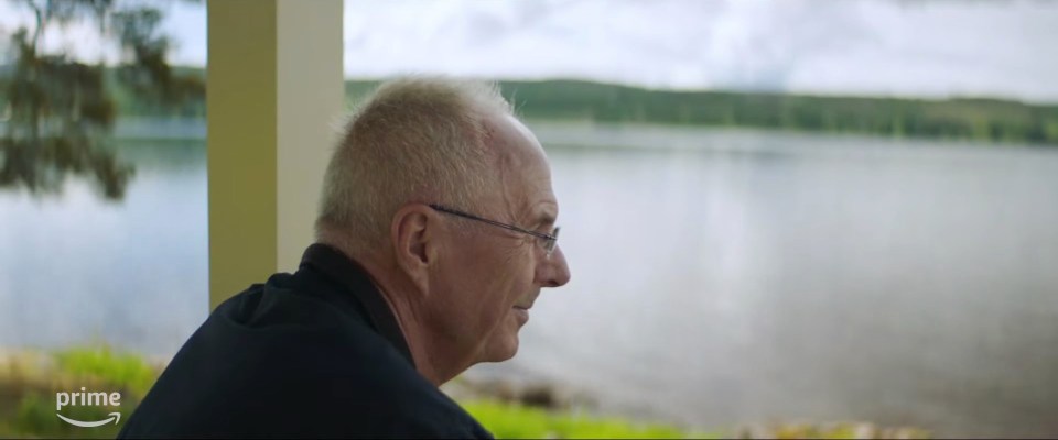 a man with glasses sits on a porch overlooking a lake