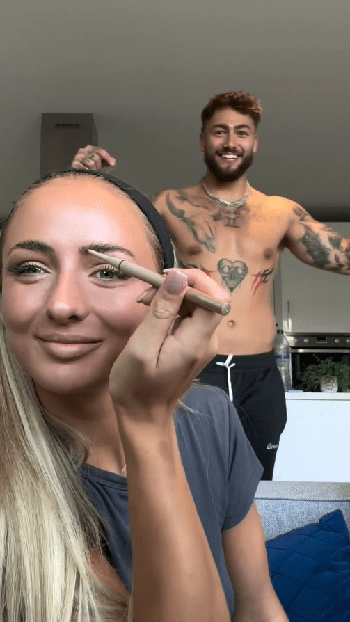 a shirtless man stands behind a woman applying makeup