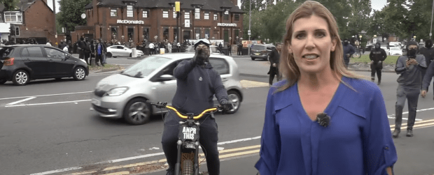 A masked protester approaches a Sky News reporter during a live report