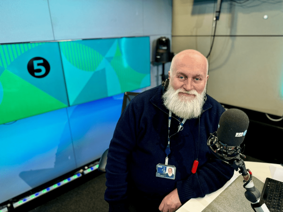 a man with a beard stands in front of a microphone with the number 5 on the wall behind him