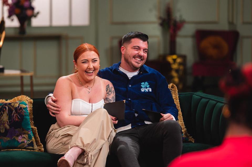 a man and a woman are sitting on a couch and the man is wearing a jacket with a clover on it