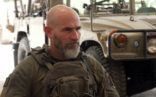 a bald man with a beard is standing in front of a military vehicle .