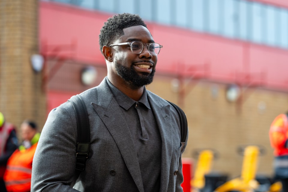 a man wearing glasses and a grey suit smiles in front of a red building