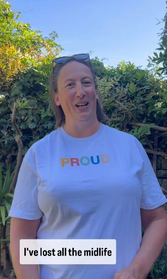 a woman wearing a white t-shirt with the word proud on it is standing in front of a bush .