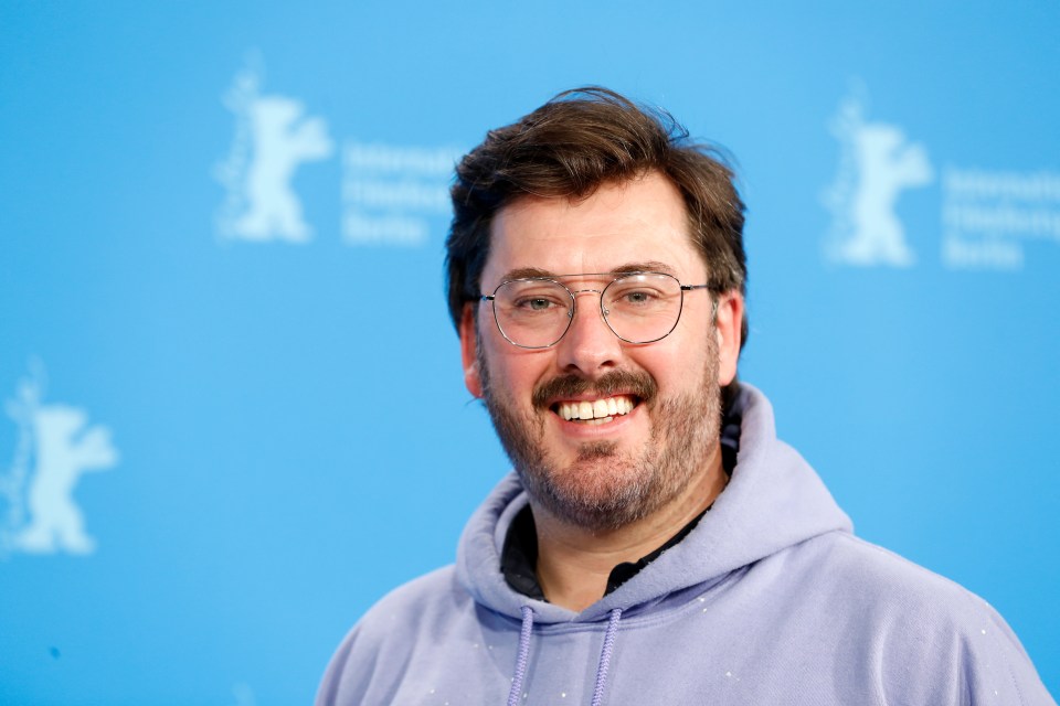 a man wearing glasses and a purple hoodie smiles for the camera