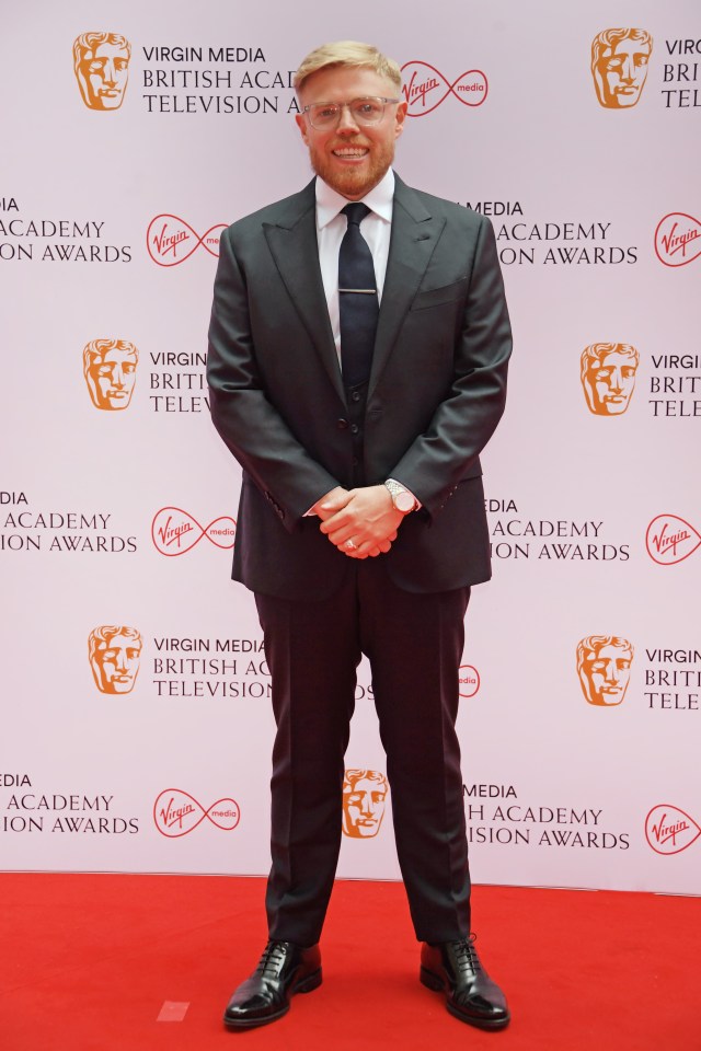 a man in a suit stands in front of a virgin media british academy television awards wall