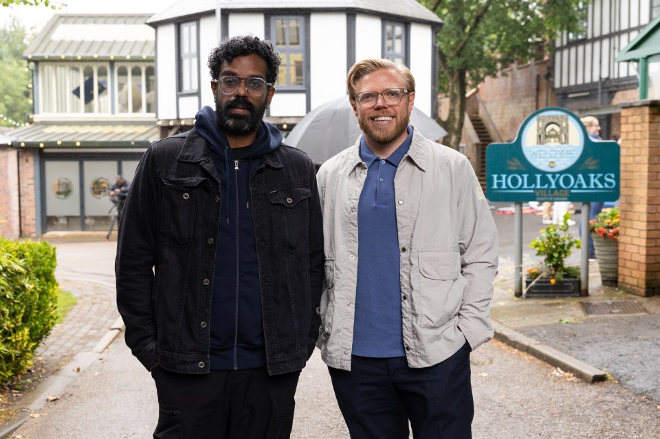 two men stand in front of a hollyoaks sign