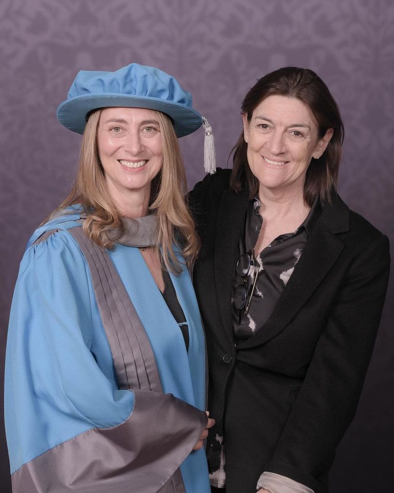 a woman in a blue cap and gown stands next to another woman in a black jacket