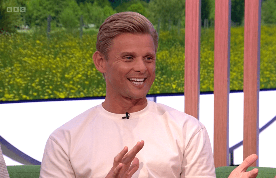 a man in a white shirt is smiling while sitting on a green couch in front of a bbc logo