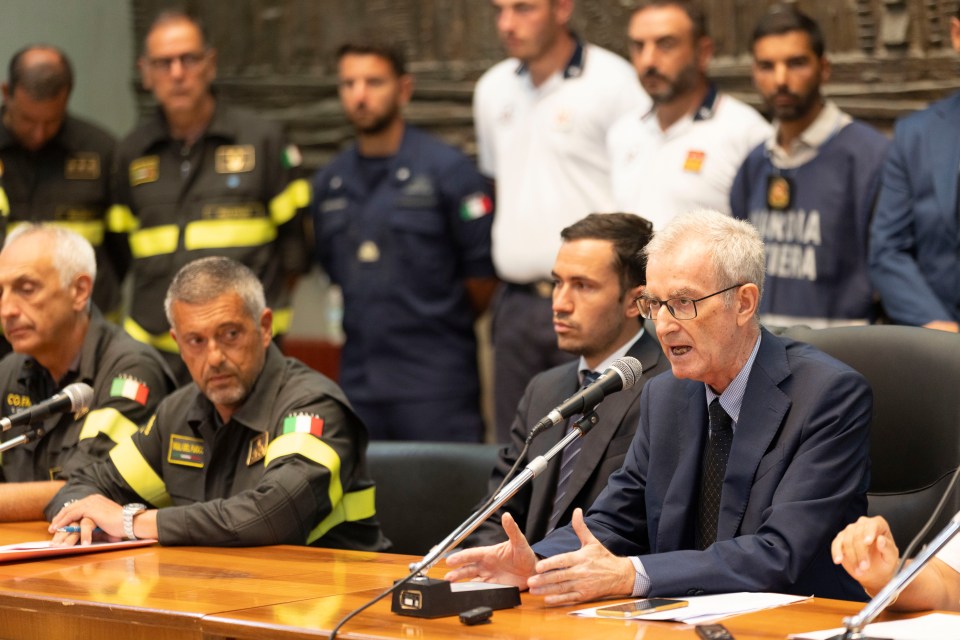 a man speaking into a microphone with a man in a marina nera shirt behind him