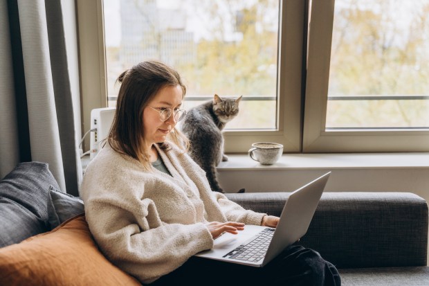 a woman sits on a couch using a laptop next to a cat