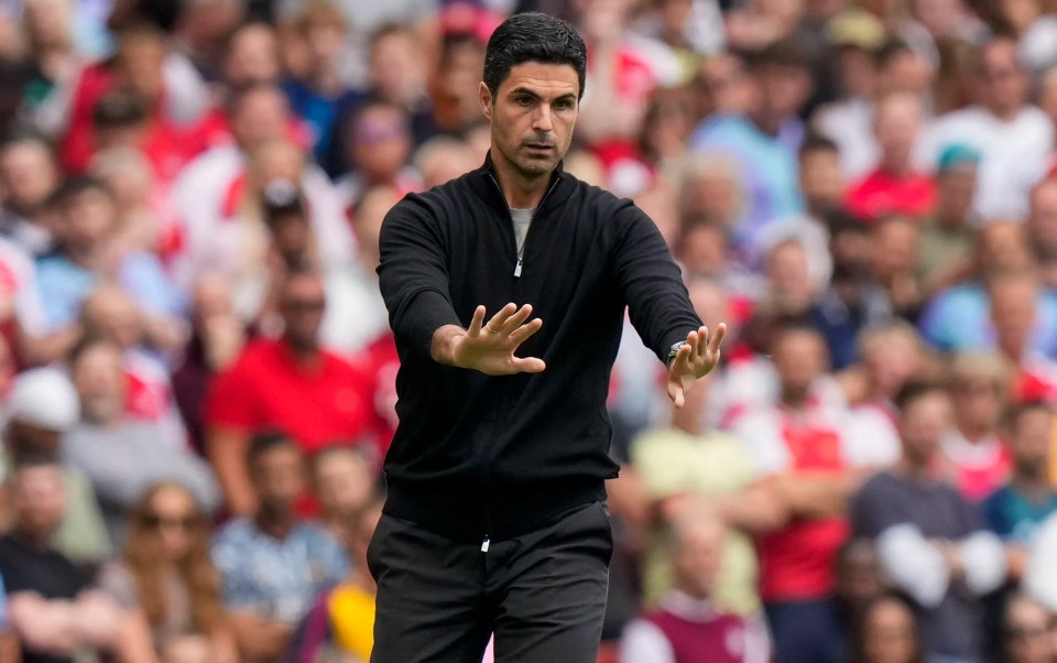 a man in a black jacket stands on a soccer field with his hands outstretched