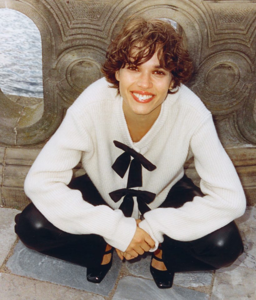 a woman in a white sweater with a black bow sits on the ground